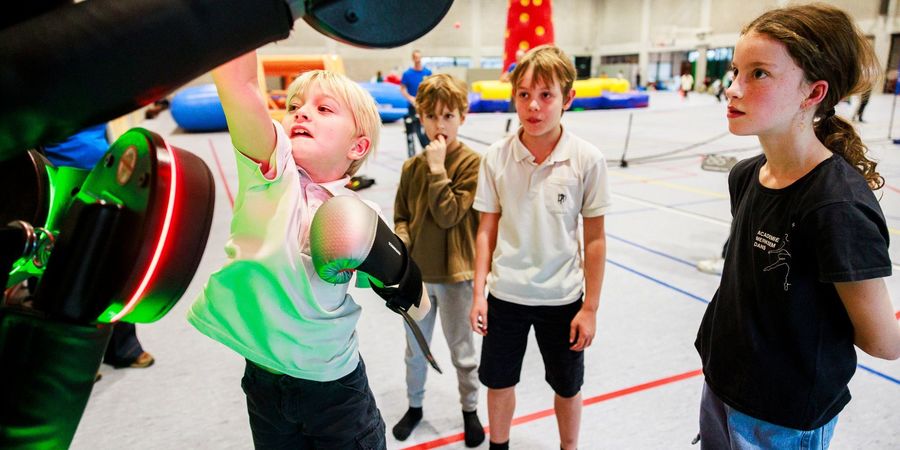 Kinderen tijdens de Fun Games in sporthal De Rode Loop. Op de voorgrond bokst een kind op een verlicht speeltoestel, terwijl andere kinderen toekijken.